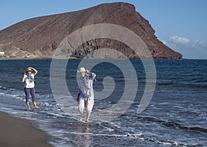 Retired senior couple walking on the shore with their feet in the water while holding their hats due to the wind. Happy active