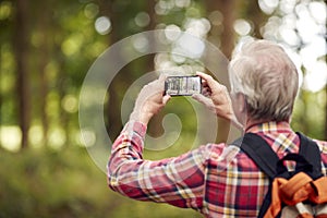 Retired Senior Man Hiking In Woodland Countryside Taking Photo With Mobile Phone