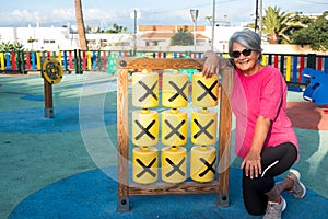 A retired senior woman with gray hair and casual clothing resting in a public park with children`s games. Sunset light