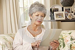 Retired Senior Woman Sitting On Sofa At Home Using Tablet Computer