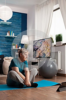 Retired senior woman in sportswear sitting on yoga mat watching fitness lesson on laptop