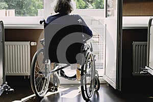 Retired senior woman on a wheel chair at hospital sadly looking looking through balcony doors outside