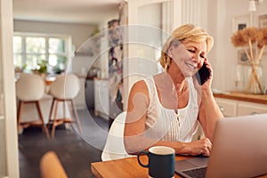 Retired Woman On Phone At Home In Kitchen Using Laptop