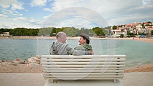 Retirement holiday, joyful elderly woman and man are sitting in an embrace on a bench near sea coast during a tourist