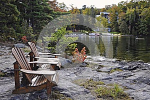 Retirement Living - Two Muskoka chairs sitting on a rocky shore facing a calm lake with trees and a white cottage in the backgroun
