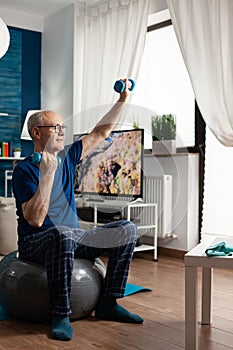 Retirement senior man sitting on aerobics swiss ball raising hand streching arm muscle