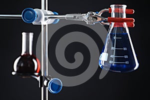 Retort stand and laboratory flasks with liquids on black background, selective focus