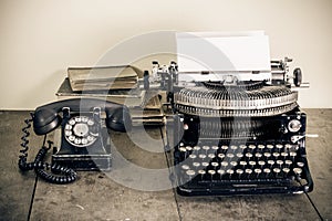 Retro aged black typewriter with paper blank,  telephone, books on wooden table. Vintage style sepia photo