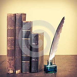 Retro aged books, quill pen and inkwell on oak wooden table. Vintage style filtered photo