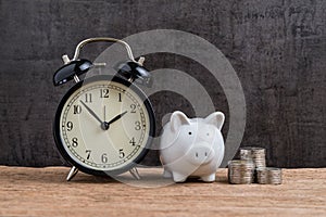 Retro alarm clock with white piggy bank and stack of coins on wooden table and black background as long term savining money or re