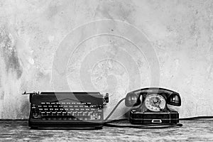 Retro black rotary telephone and typewriter on wooden table in front gray concrete background