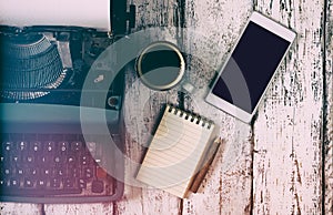 Retro filtered image of vintage typewriter, blank notebook, cup of coffee and smartphone on wooden table