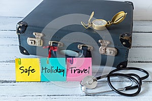 retro style suitcase with compass and medical stethoscope. Colorful sticky notes with the words `Safe, Holiday, Travel`. The conc