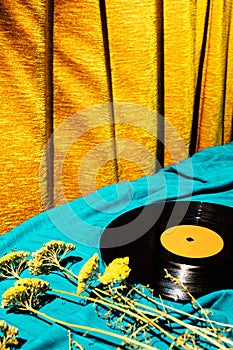 Retro styled image of LP vinyl record on a cyan silk or saten fabric with dry old flowers and gold orange curtain in the