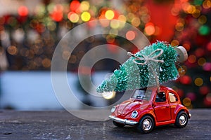 Retro toy red car carrying tiny Christmas tree on the background of christmas bokeh. Christmas cards. Trucking.Delivery service.