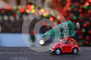 Retro toy red car carrying tiny Christmas tree on the background of christmas bokeh. Christmas cards. Trucking.Delivery service.