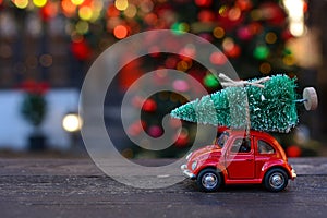 Retro toy red car carrying tiny Christmas tree on the background of christmas bokeh. Christmas cards. Trucking.Delivery service.