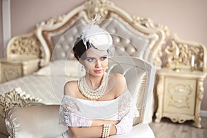 Retro woman portrait. Elegant brunette lady in hat with hairstyle, pearls jewelry set. Pretty female posing on modern armchair in