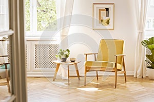 A retro, yellow armchair and a wooden table in a beautiful, sunny living room interior with herringbone floor and white walls. Re