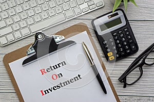 RETURN ON INVESTMENT text with notepad, keyboard, decorative vase, fountain pen, spectacles and calculator on wooden background