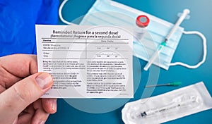 Return for a second dose card with facial mask, covid-19 vaccine vial and syringe on blue