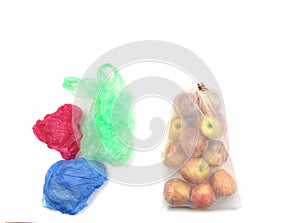 Reusable recycled mesh net grocery bag full of apples and heap of colored disposable plastic bags, on white background, benefit