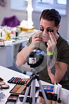 Curious extraordinary man sitting at the table surrounded with makeup