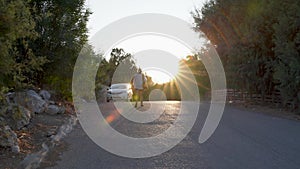Rhodes, Greece: A young girl in white dress walking towards car vehicle during sunset / sun rise on a steep road in a middle of a