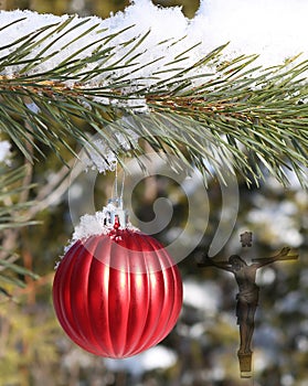 Ribbed red ball hanging from outdoor evergreen tree in the snow with semi-transparent cross and body of Jesus