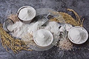 Rice flour Thai organic in a measuring spoon with wooden bowl on a pile of white rice, ear of paddy and strainer on concrete