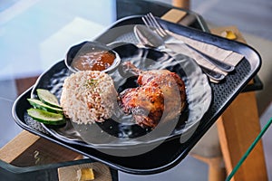 Rice with grilled chicken leg and cucumber on black plate in a tray on a glass table