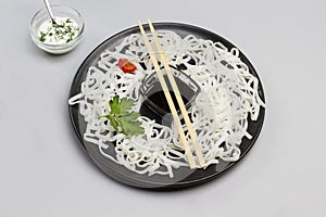 Rice noodles with sauce, bamboo sticks on black plate. Cherry tomatoes in bowl with white sauce on table