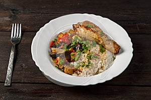 Rice with stewed vegetables in sauce and chicken leg in a white plate on a wooden table