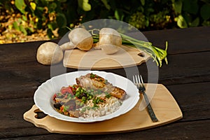 Rice with vegetables and chicken leg on a handmade wooden board next to champignons and green onions on a wooden table against a