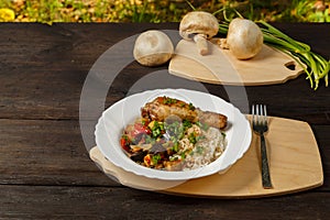 Rice with vegetables and chicken leg on a handmade wooden board next to mushrooms and green onions on a wooden table