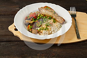 Rice with vegetables and chicken leg in a white plate on a handmade wooden board next to a fork on a wooden table