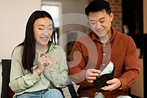 Rich asian mature husband giving money to his wife, sitting on sofa at home. Financial abundance family budget concept