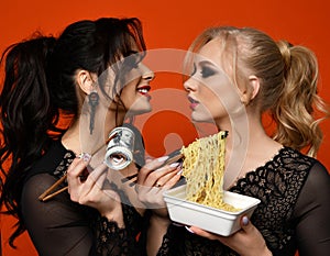 Rich brunette women holds stack of money cash with chopsticks and looks at at her poor blonde girl eats cheap noodles