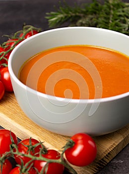Rich color tomato puree soup in a gray bowl on a wooden board with cherry tomatoes and herbs on a background