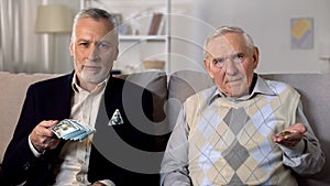 Rich elderly man showing bunch of dollars, poor senior friend showing coins photo