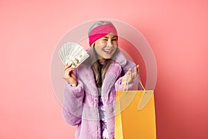 Rich and fashionable asian senior woman wasting money in shops, holding shopping bag and dollars, winking happy at