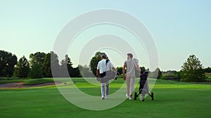 Rich golf players walking on field. Two active sportsmen carry equipment clubs.