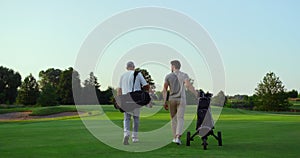 Rich golf players walking on field. Two active sportsmen carry equipment clubs.