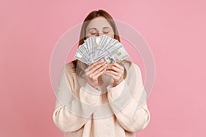 Rich greedy woman holding big fan of dollars bills and smelling money with satisfied expression.