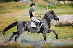 Rider man galloping fast in water pond on black stallion horse during eventing cross country competition
