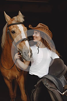 Portrait of rider woman in a leather hat with a brown horse in a stable