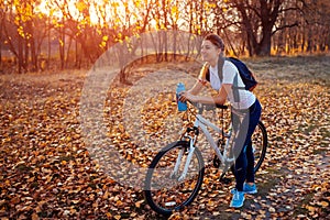 Riding bicycle in autumn forest. Young woman having rest after workout on bike holding water bottle. Healthy lifestyle