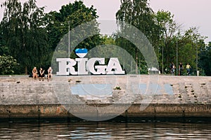 Riga Latvia. Three Young Women Sitting On Edge Of Concrete Embankment Of Daugava River