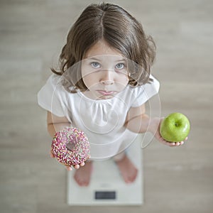 Right choice. Little sad girl holding in hands green apple and highcalorie donut. Kid trying to make decision between healthy