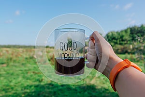 Right hand of woman holding cup of black hot Americano coffee. Clear glass with words GOOD MORNING on a green field background
